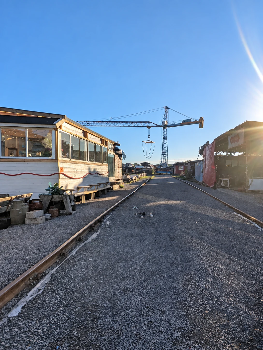 Räls mellan varvets byggnader med en stor kran under blå himmel.
