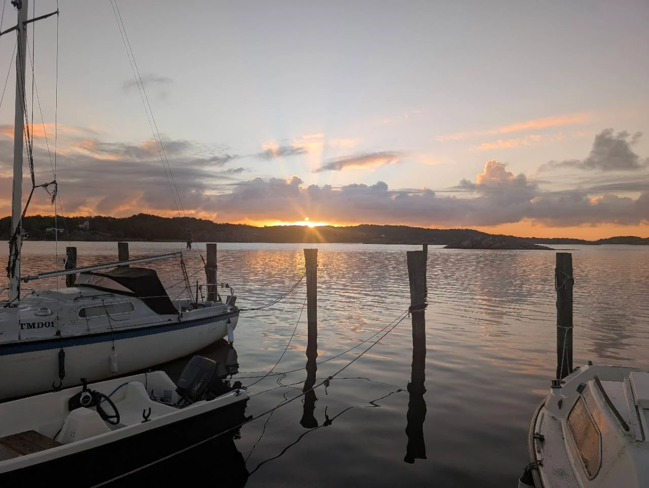 Solen vid horisonten över stilla vatten, med båtar och bryggpålar i förgrunden.